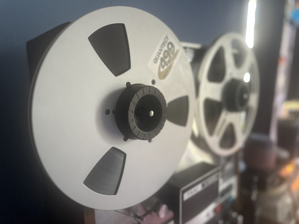 Side view of a Quantegy 499 Grand Master Gold reel on a TEAC A-6300 tape machine, highlighting analog recording equipment used in full music production. Emphasizes high-fidelity audio recording for professional sound engineering.